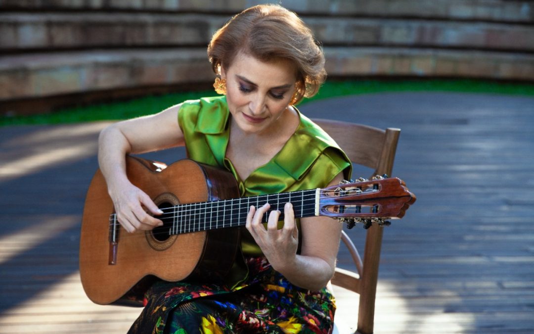 Berta Rojas tocando la guitarra sentada en una silla con un vestido floreado